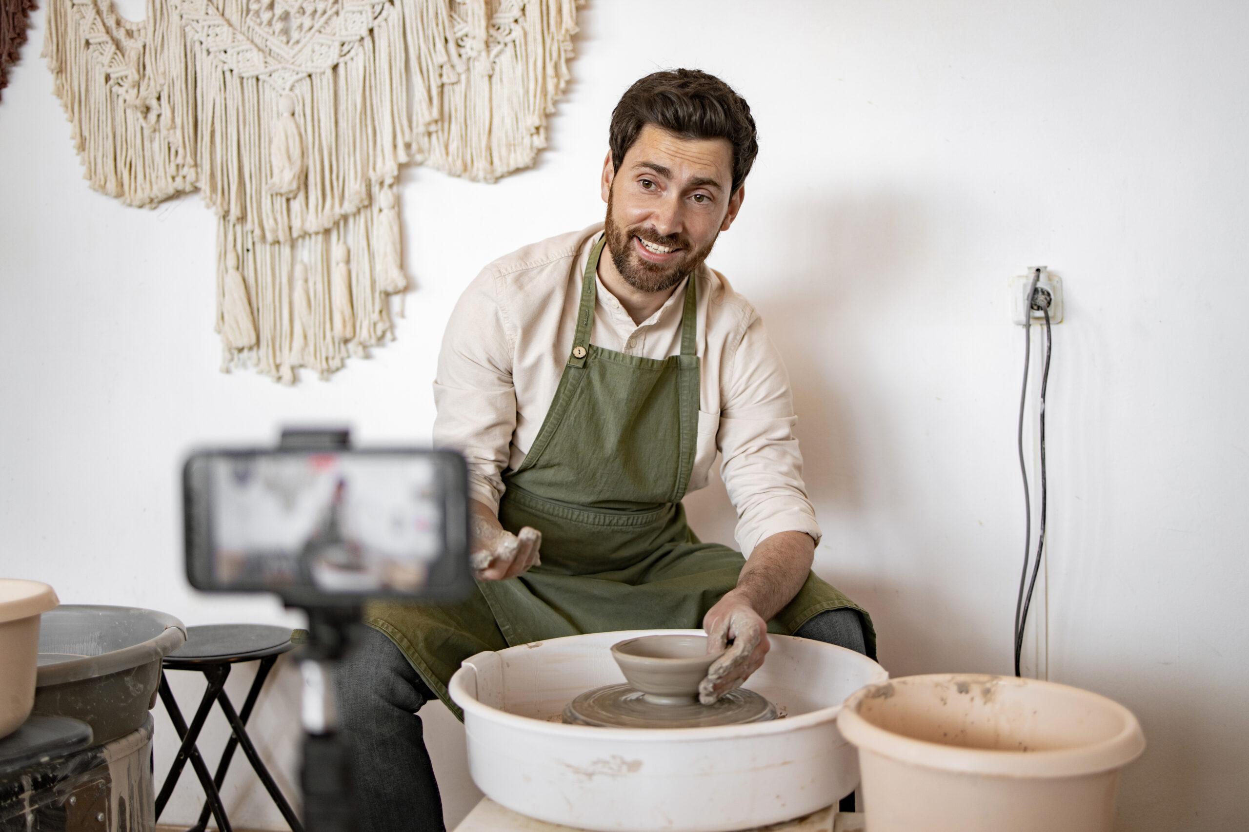 Young male potter sculpts a vase on a potter's wheel while recording a video blog using a smartphone. Creative activity and content creation.